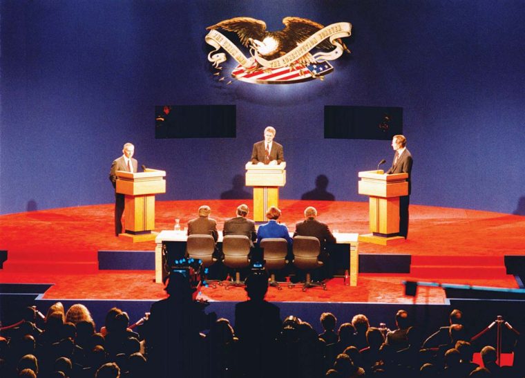 1992 presidential debate stage. Ross Perot, Bill Clinton and George HW Bush stand behind podiums on a red carpeted stage.