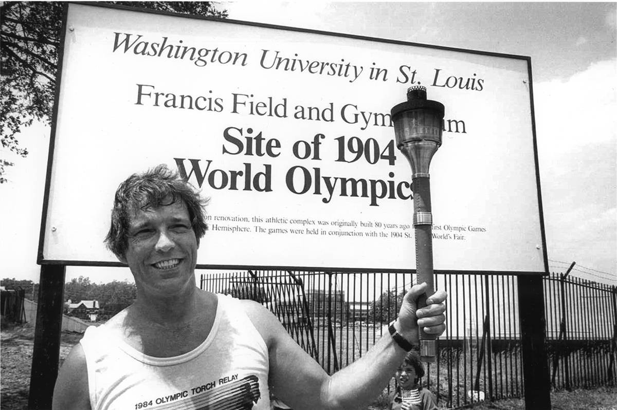 man carries Olympic torch in front of Francis Field sign