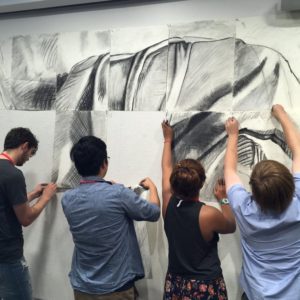 four students hang pieces of a charcoal drawing on a wall