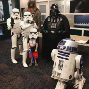 four adults dressed as stormtroopers, Darth Vader and Chewbacca pose with a boy in a stormtrooper helmet. R2D2 figure in foreground.