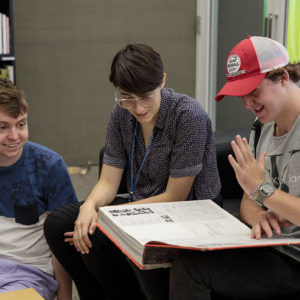three male students smile over a large book
