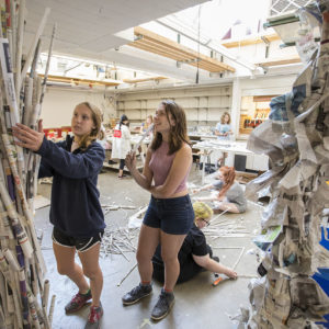 two women in shorts and sneakers affix pieces of rolled-up newspaper to a ceiling-high newspaper sculpture.