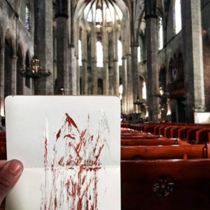a hand holds a sketchbook with a rough drawing in red ink up to the inside of a cathedral, which it depicts.