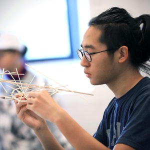 a young man with a short black ponytail and glasses holds a delicate structure constructed of tiny wooden dowels.