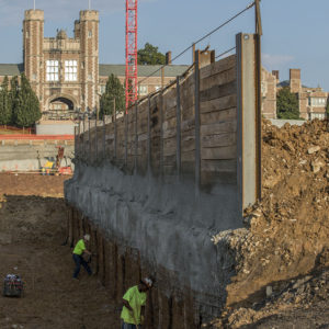 two construction workers work in a trench below the beginning of a wall. Brookings Hall in the background.