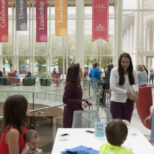 people gather around a table and chat in a large glass atrium. Behind them are banners with values printed on them: Diversity, Excellence, etc.