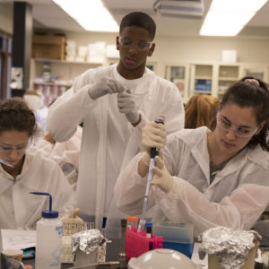 Two young women work with pipettes at a lab table, wearing safety glasses and white coats. A young man stands behind them holding other materials.