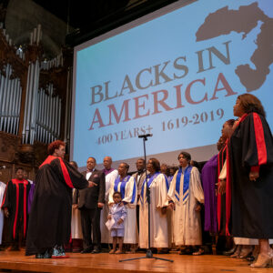 Gospel choir stands while singing on stage with Blacks in America slogan projected behind.