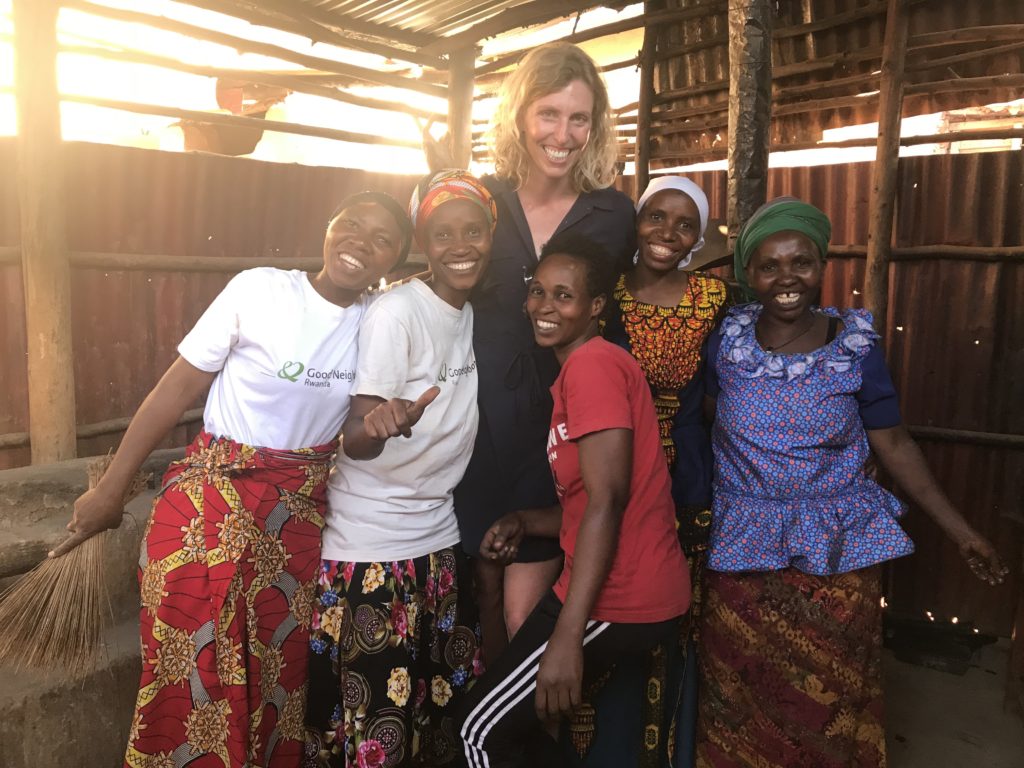A tall Caucasian woman and a group of East-African women stand smiling inside a makeshift building