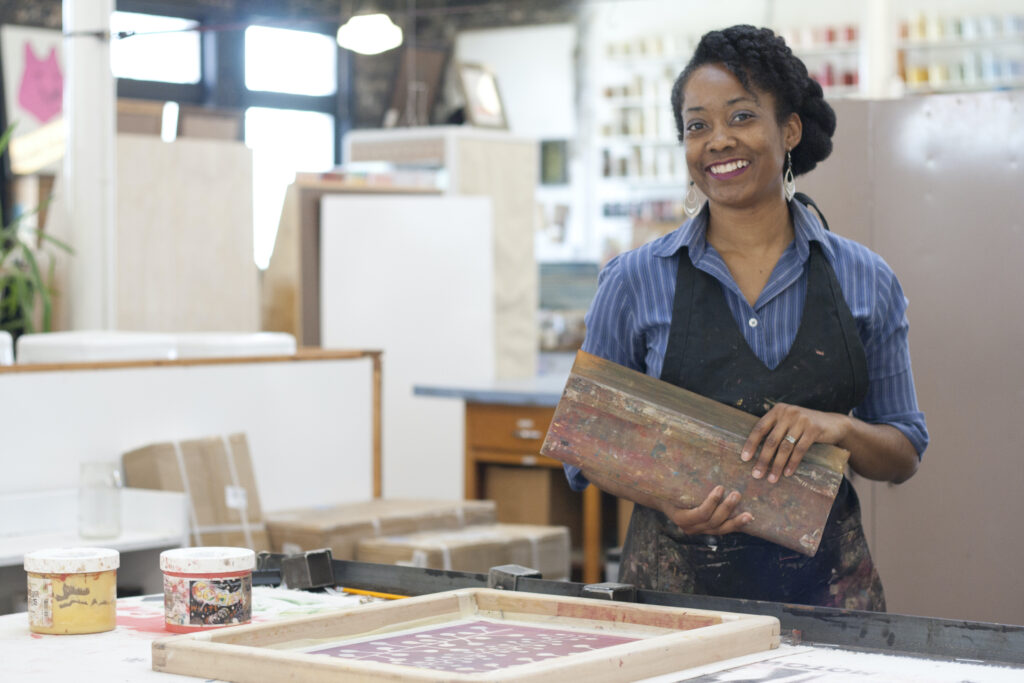 African American woman artists stands in a screen printing studio holding her own work 