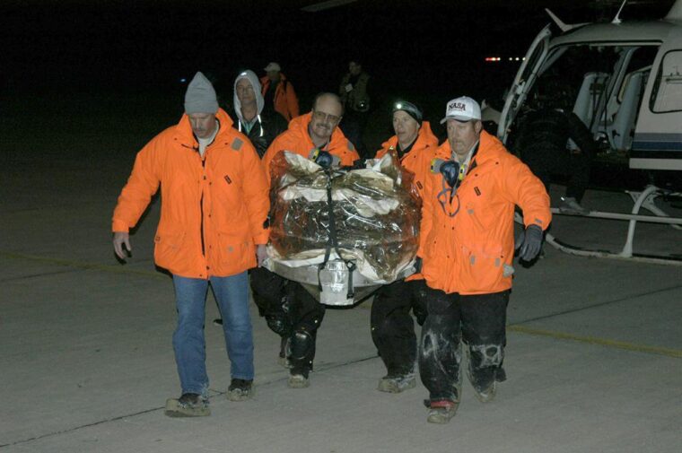 Workers in orange coats carry the Stardust mission return capsule inside a protective covering.