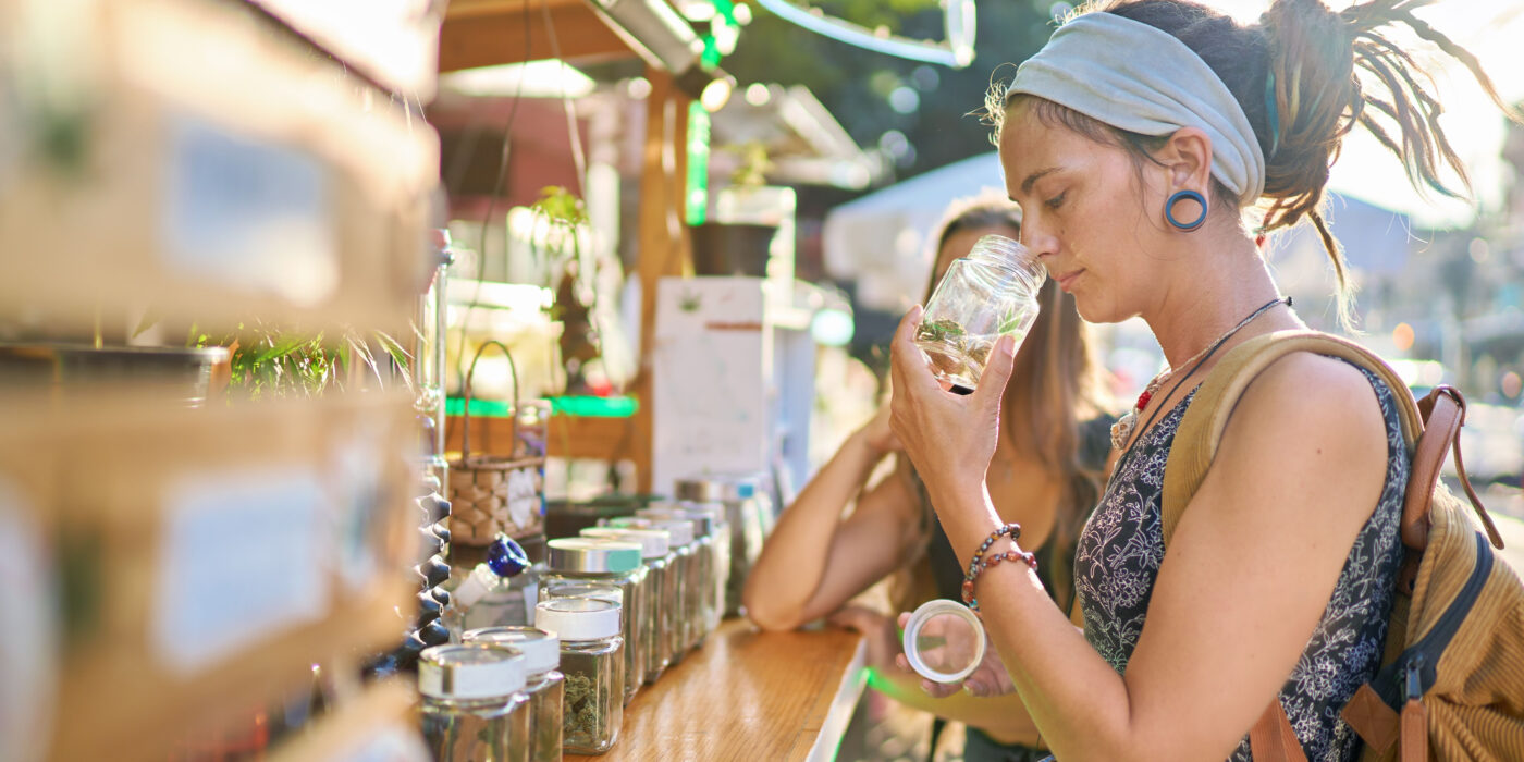 A white woman with dreads and a headband sniffs from a jar of cannabis